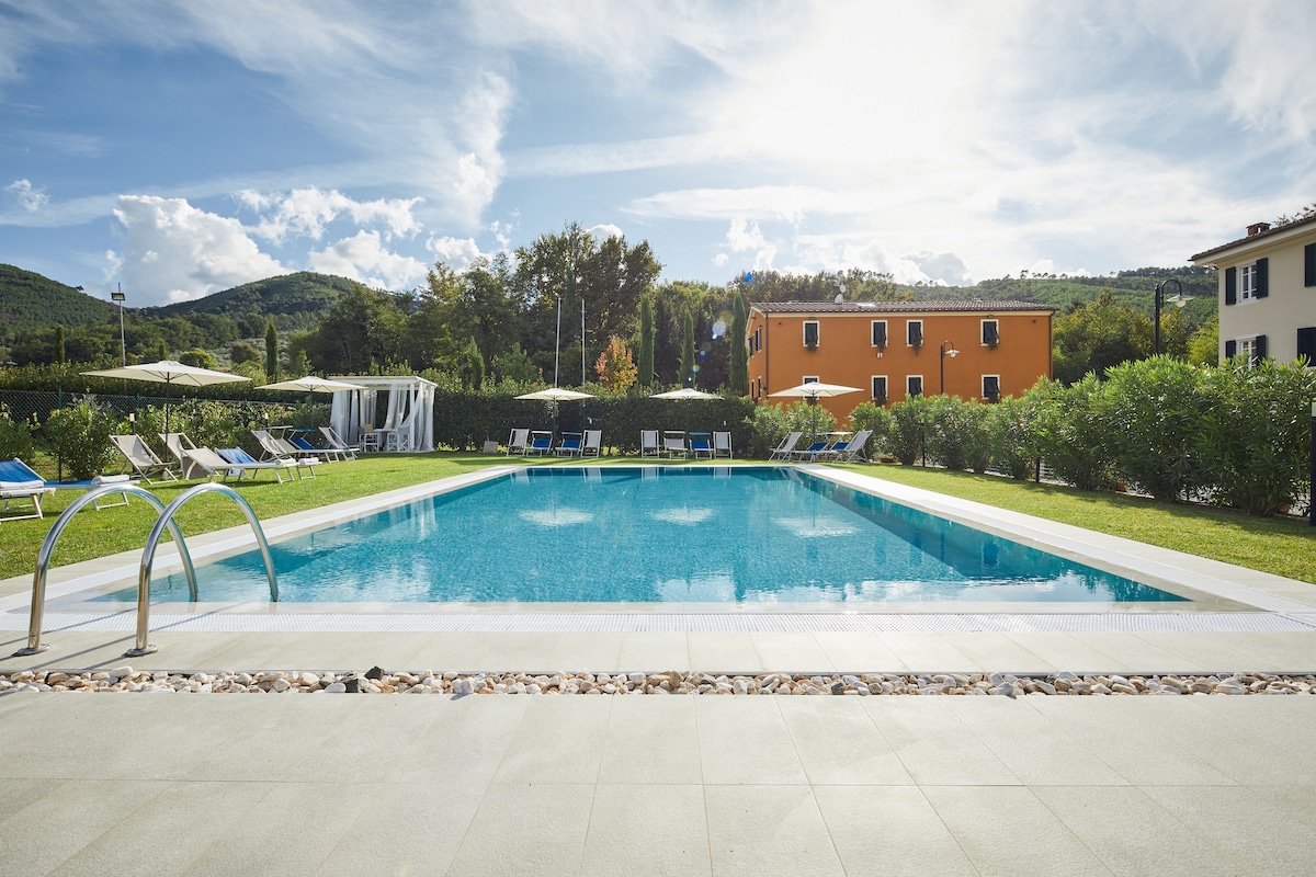 A tranquil pool area is framed by manicured grass and surrounded by olive trees. Sun loungers line the poolside, and umbrellas provide shade. The vibrant orange building in the background complements the natural landscape, while the sky is partly cloudy, allowing sunlight to reflect on the water.