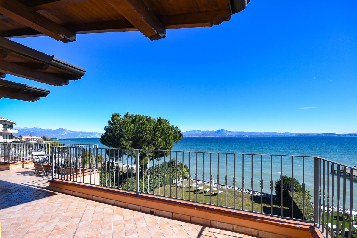 A spacious terrace offers a panoramic view of the lake and surrounding landscape. The clear blue water contrasts with the vibrant greenery and distant mountains. Comfortable seating options are visible on the terrace, creating an inviting outdoor space.