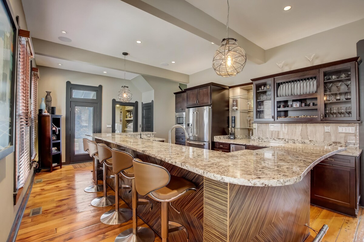 A modern kitchen is presented with a large granite countertop and six bar stools for seating. Sleek cabinetry and glass shelves display dinnerware, while stainless steel appliances are integrated into the design. Soft lighting is provided by pendant fixtures, and natural light enters through nearby windows.