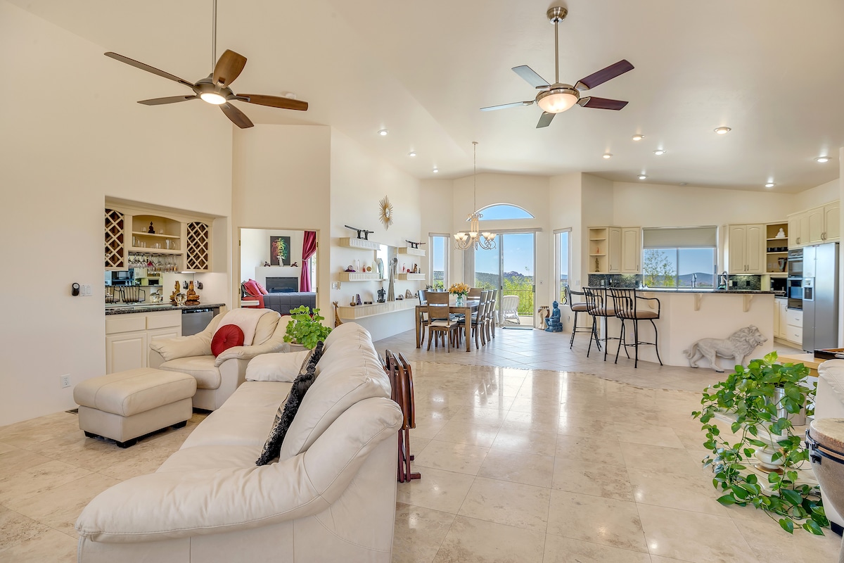 An open living space features vaulted ceilings with natural light flooding through expansive windows. A spacious sectional sofa is positioned in front of a sleek coffee table, while a dining area and kitchen are visible in the background, accentuated by ceiling fans and light-colored cabinetry.