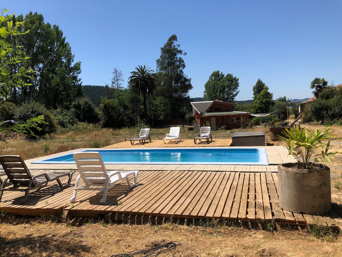 A swimming pool is positioned on a wooden deck surrounded by sun loungers. Lush greenery and trees provide a natural backdrop, while a glimpse of the apartment is visible in the distance. The tranquil setting creates an inviting space for relaxation.