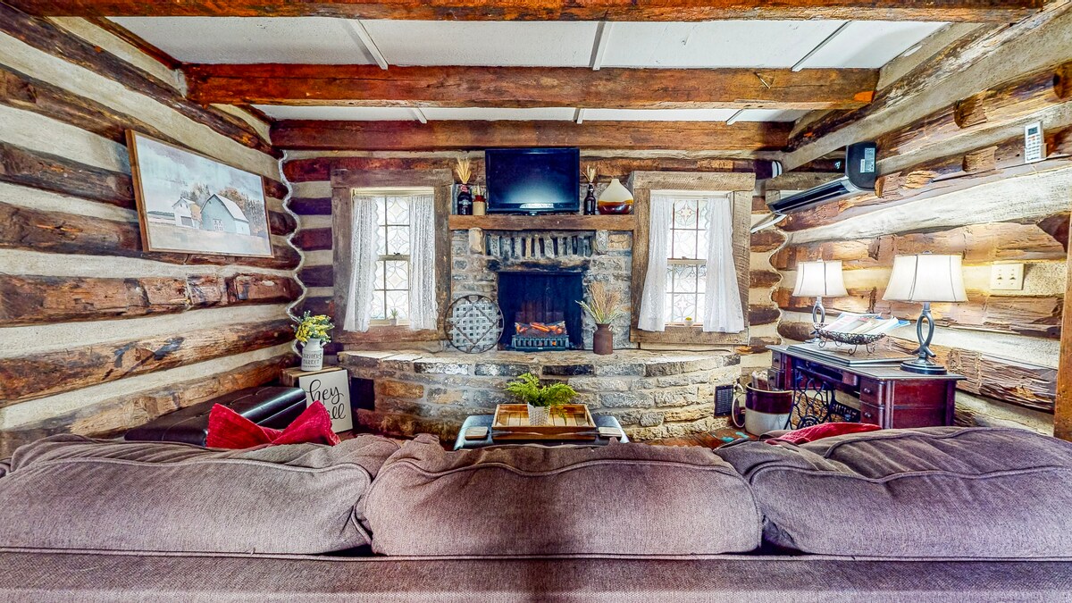 A cozy living room features rustic log walls and a stone fireplace with an electric insert. Two windows allow natural light to illuminate the space. A comfortable sofa, adorned with throw pillows, faces the fireplace, while a wooden coffee table and side lamps add warmth.