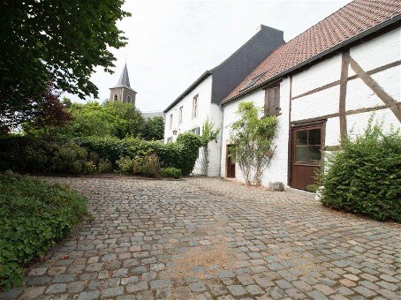 A cobblestone courtyard is framed by lush greenery and various shrubs, providing a serene setting. The historic building features a mix of whitewashed walls and wooden beams, with a spire from a nearby church visible in the background, adding character to the peaceful environment.