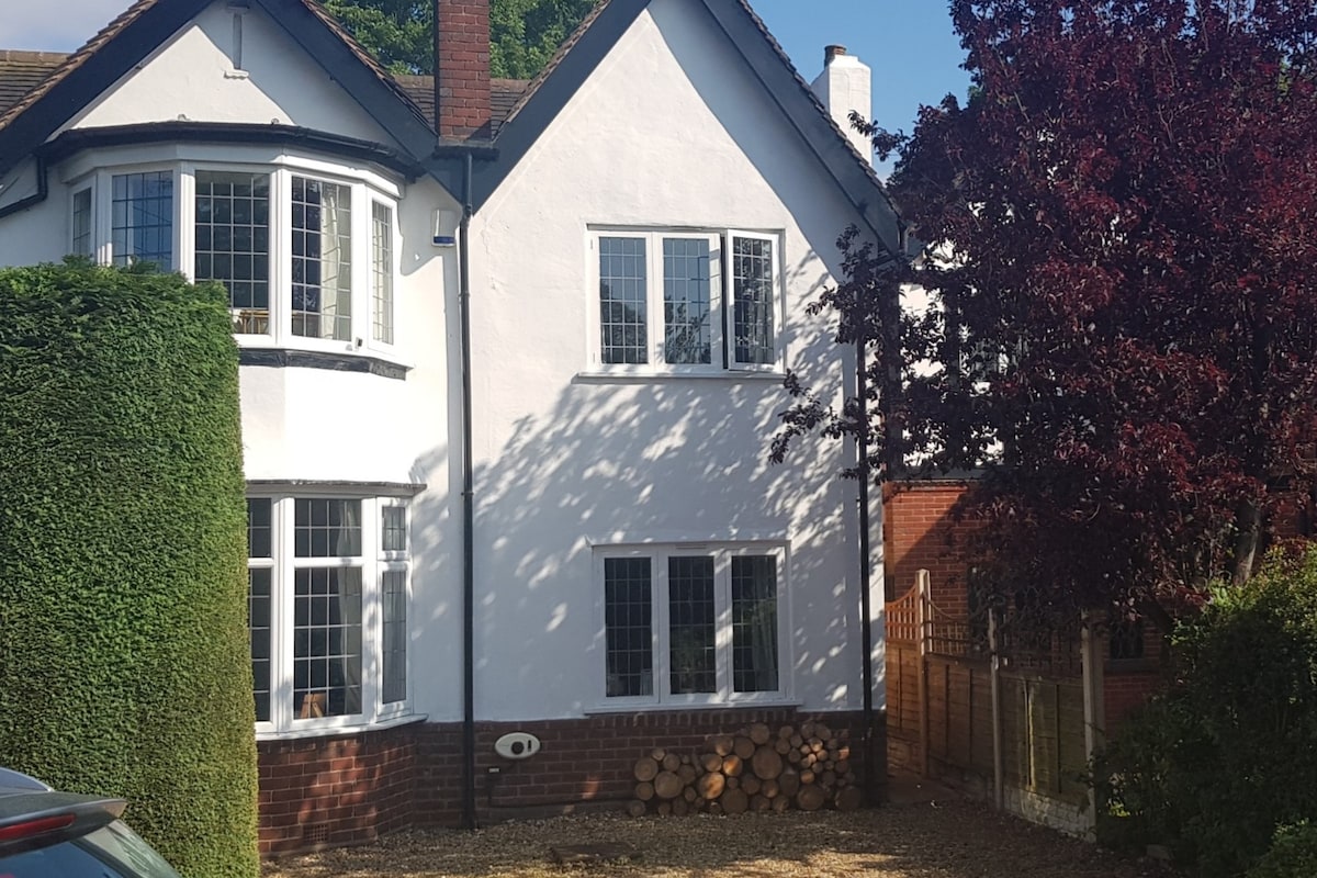 The exterior of a well-maintained, two-story house is visible, featuring white walls and large front windows. A manicured hedge frames the property, with a stack of logs neatly arranged against a wooden fence in the background.
