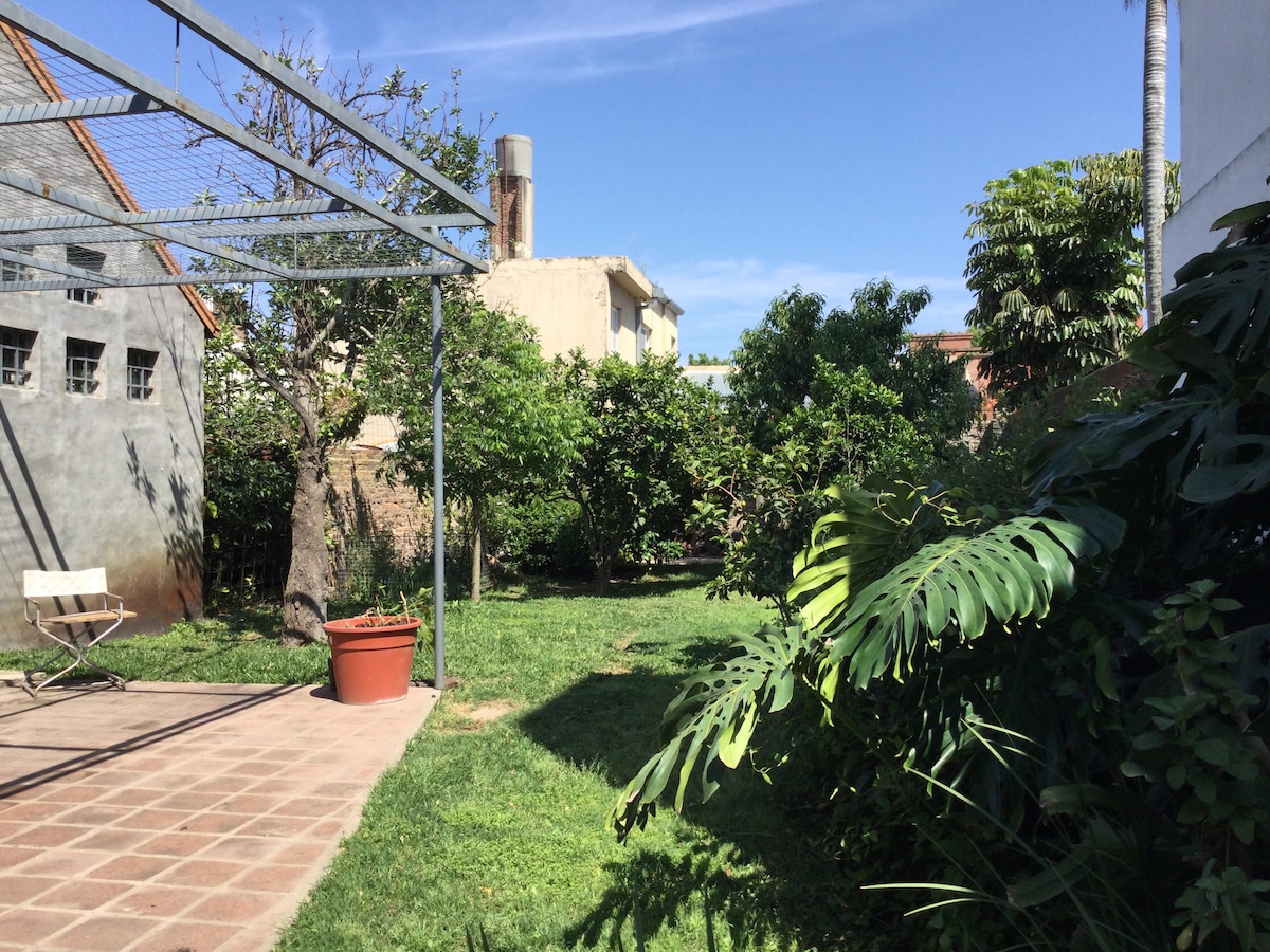 A spacious garden area is visible, featuring a variety of lush green plants and trees. A canopy frame suggests a shaded space, while a paved section with a single chair enhances outdoor relaxation. The bright sky and greenery create a serene environment.
