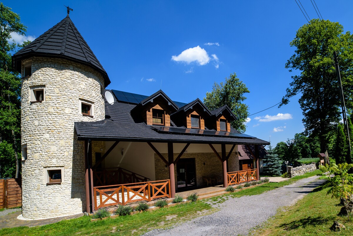 The exterior of a castle-style guesthouse features a circular stone tower with a black roof, adjacent to a modern two-level building. A wooden balcony extends from the front, while the surrounding greenery and blue sky enhance the inviting atmosphere.