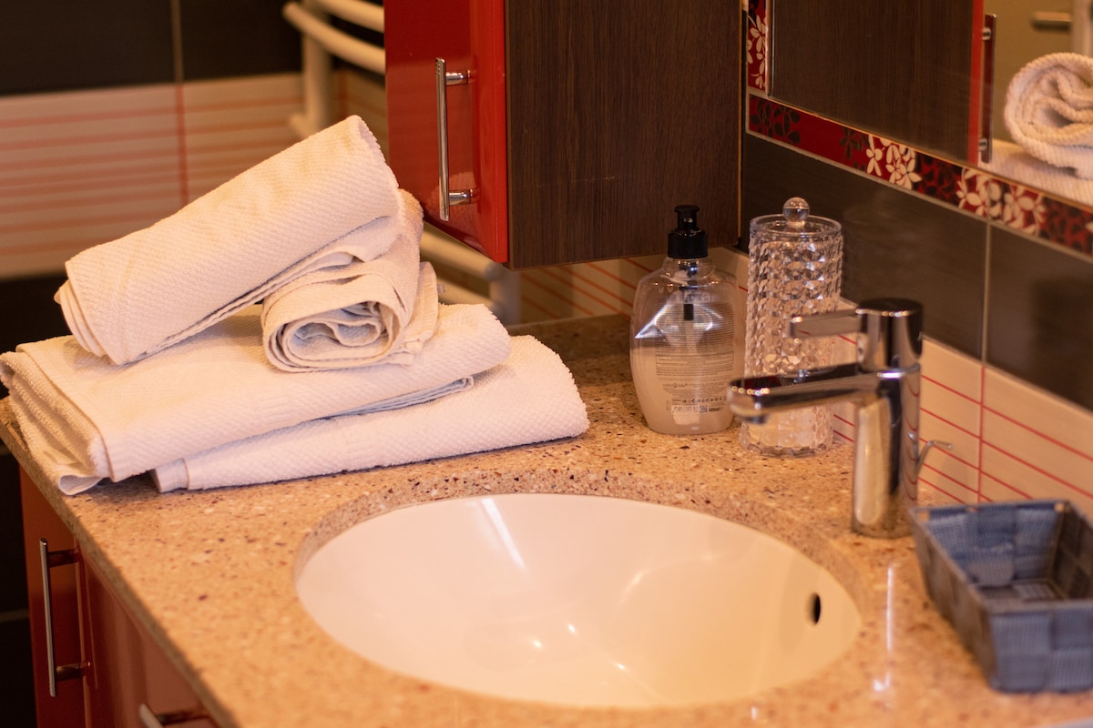 A modern bathroom countertop is featured, showcasing neatly stacked towels beside a round sink. A soap dispenser and a decorative glass container are positioned on the surface, while a small tray holds additional items, creating a tidy and functional space.