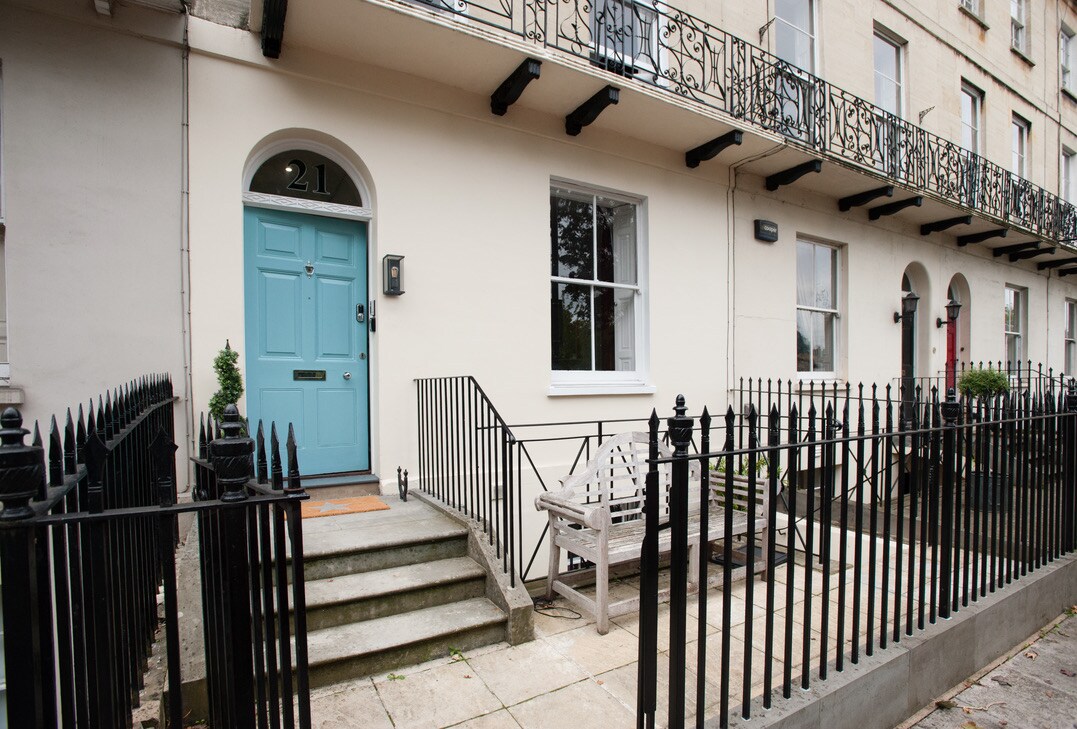 The exterior of a Georgian terrace building features a pale blue front door, flanked by a black wrought iron railing and a small set of steps. A wooden bench is positioned on the patio area, enhancing the welcoming entrance.