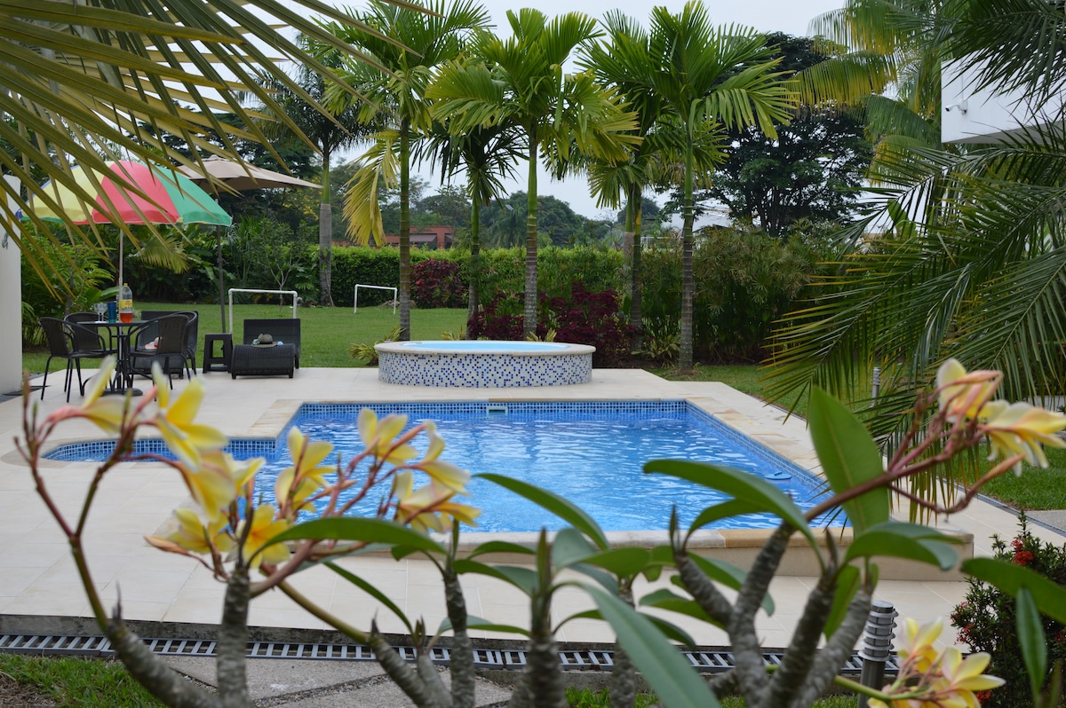 A private pool is surrounded by lush greenery, featuring comfortable seating options nearby. A jacuzzi rests at one end of the pool, while vibrant flowers frame the view, adding a touch of natural beauty. Multiple palm trees provide shade and enhance the tropical setting.
