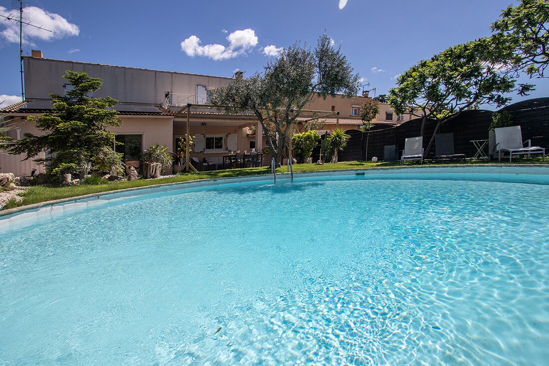 A round private pool of 50m2 is captured, offering clear turquoise water under a bright sky. The villa's outdoor space is visible in the background, surrounded by trees and well-maintained greenery, creating a serene environment for relaxation.