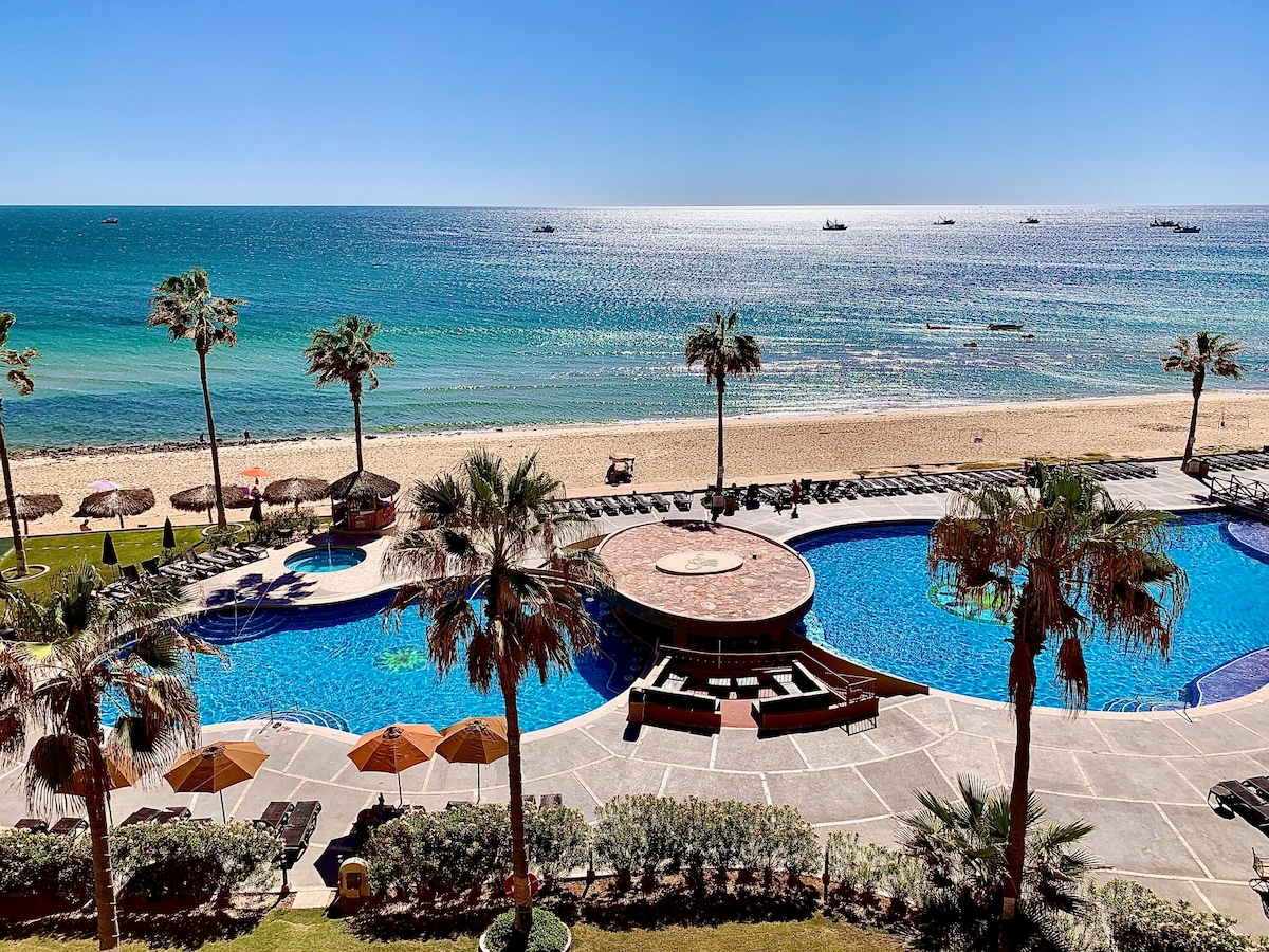 The image features a serene beachfront view with a pristine sandy beach and calm turquoise waters. Two swimming pools are visible, surrounded by palm trees and lounge chairs. Umbrellas provide shaded seating near the water's edge, enhancing the inviting outdoor atmosphere.
