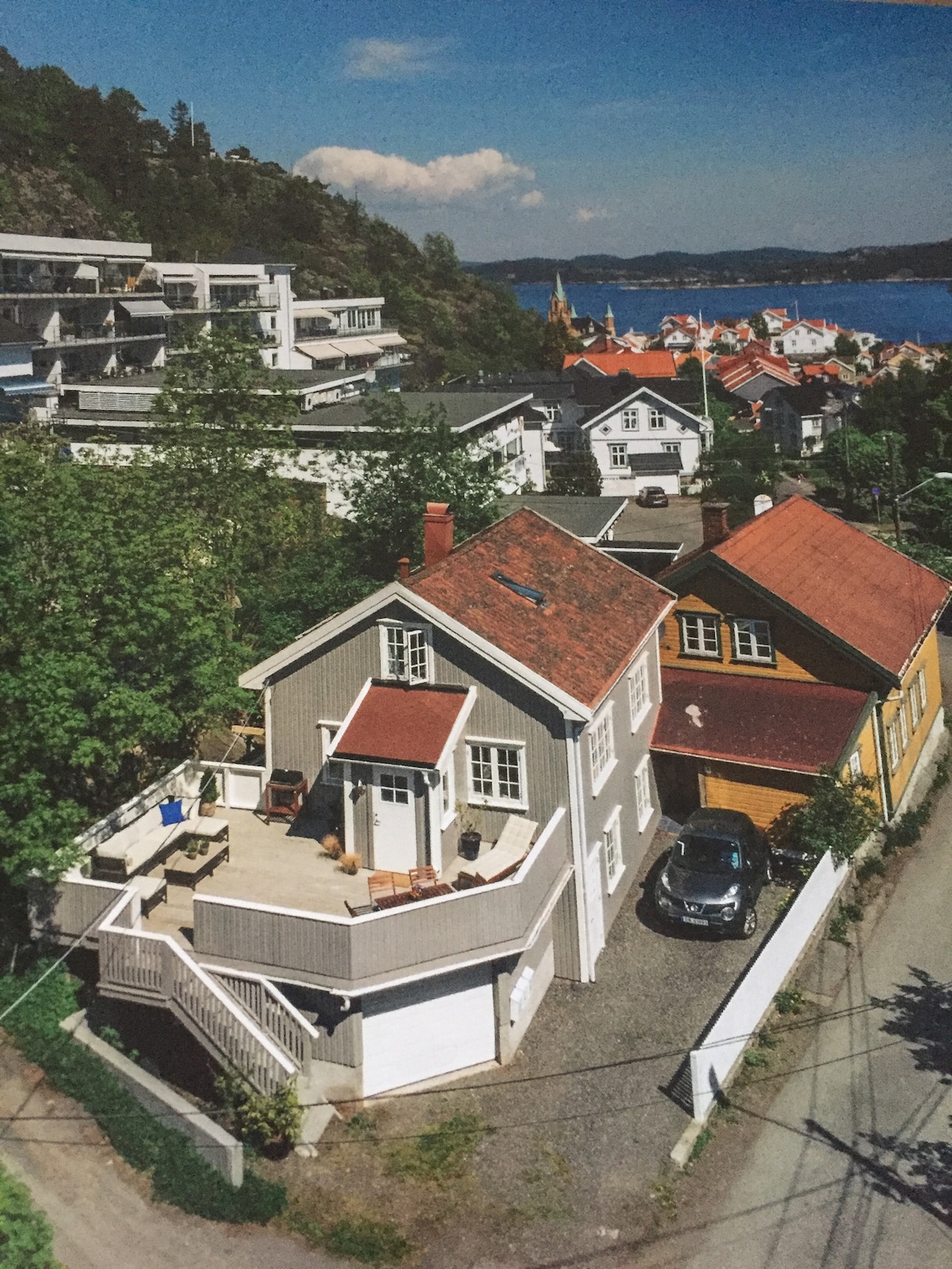 An aerial view of a charming two-story house with a red roof, situated near the coastline. A patio is visible on the upper level, furnished with outdoor seating. Surrounding houses and greenery create a cozy neighborhood atmosphere, with water in the background.