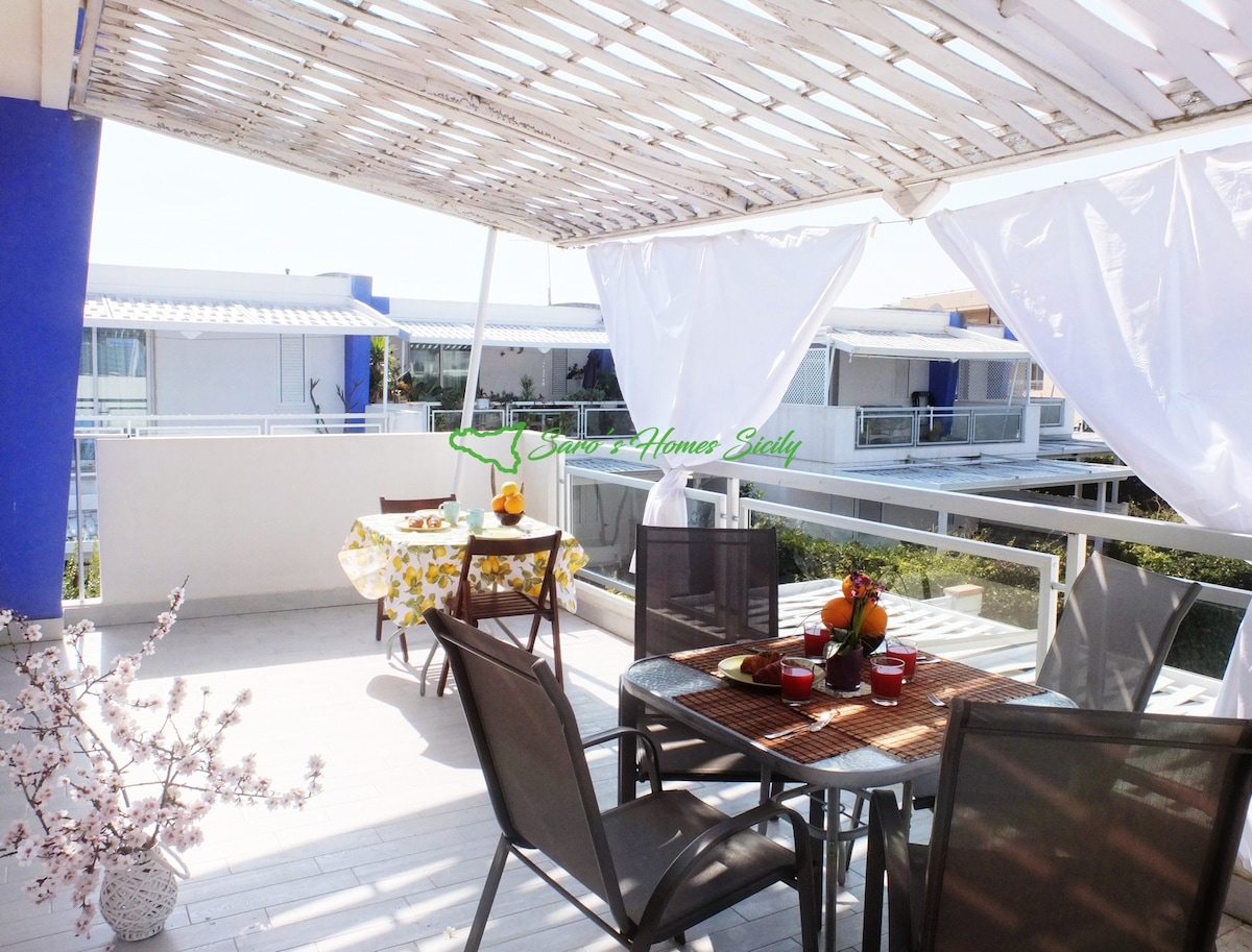 An outdoor veranda features a table set for dining, surrounded by chairs. A canopy provides shade, and decorative elements include a flowering plant. The area invites relaxation with views of the surrounding residences.