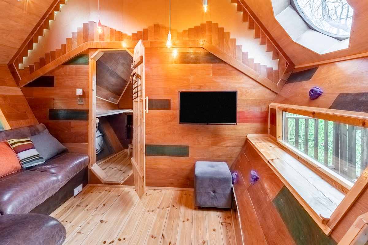 A cozy living room space features warm wooden walls and floors, complemented by a plush sofa and a small ottoman. A smart TV is mounted on the wall. A large window provides natural light and a view of the surrounding trees.