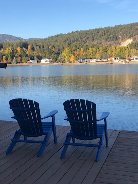 Two blue Adirondack chairs are positioned on a wooden deck overlooking the calm waters of the Pend Oreille River. The surrounding landscape features vibrant autumn foliage and distant tree-covered hills under a clear blue sky.