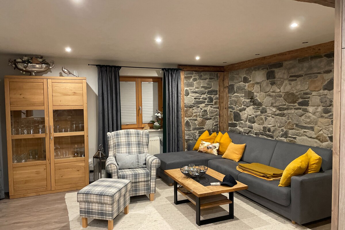 A cozy living area features a grey sectional sofa with yellow cushions, accompanied by a plaid armchair. An oak coffee table sits atop a light rug, with decorative items placed on it. A glass-front cabinet adds storage and elegance, while stone accents create a rustic charm.