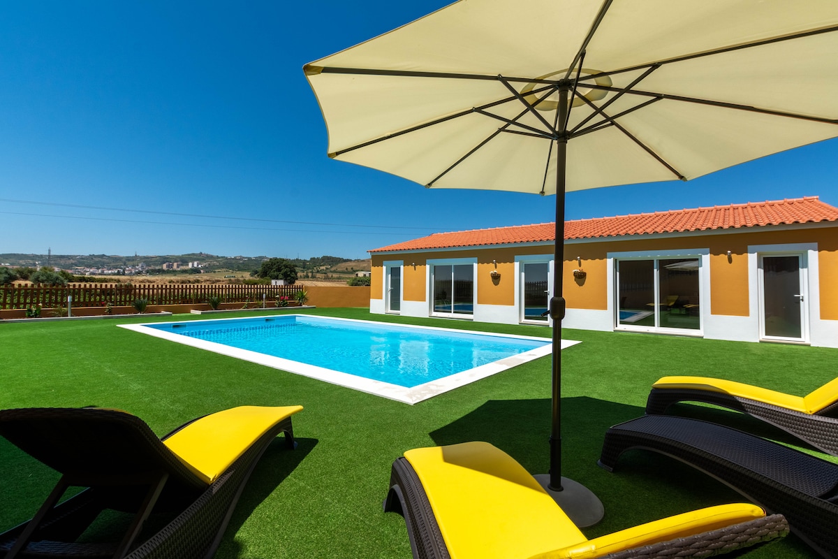 A vibrant outdoor area is presented, featuring a rectangular pool surrounded by a well-manicured lawn. Yellow lounge chairs are positioned beside the pool, shaded by a large umbrella. The background showcases a single-story building with large glass doors offering views of the landscape.