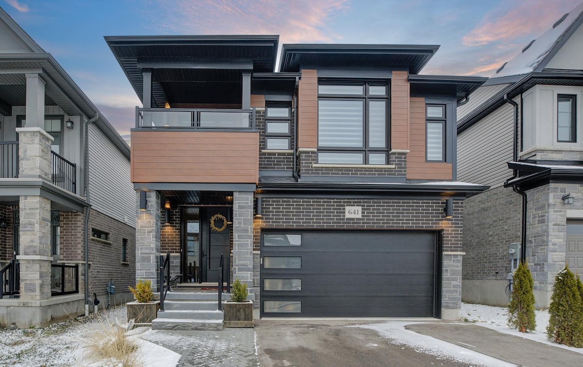 The contemporary exterior of a two-story home is highlighted, featuring a combination of sleek brick and wooden accents. A spacious driveway leads to a modern garage door. A small pathway bordered by minimal landscaping leads to the entrance, framed by wide pillars and large windows.
