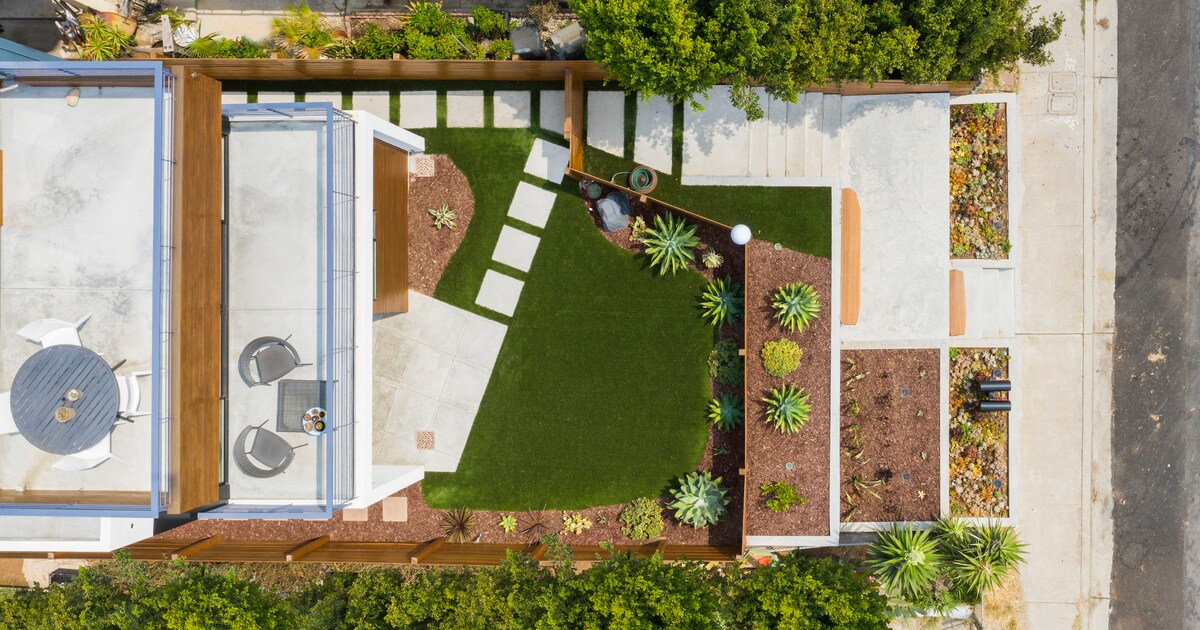 An aerial view captures a well-maintained semi-private front yard, featuring patches of green grass and landscaped areas with various plants. A stone pathway leads through the space, while a small seating arrangement is visible on a concrete patio. The yard is bordered by wooden fences.