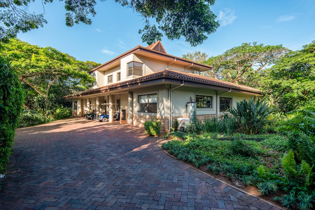 A two-story house is presented with a broad driveway bordered by lush greenery. Large windows and a sloped roof create a welcoming façade, while outdoor seating areas offer a connection to the surrounding natural landscape.
