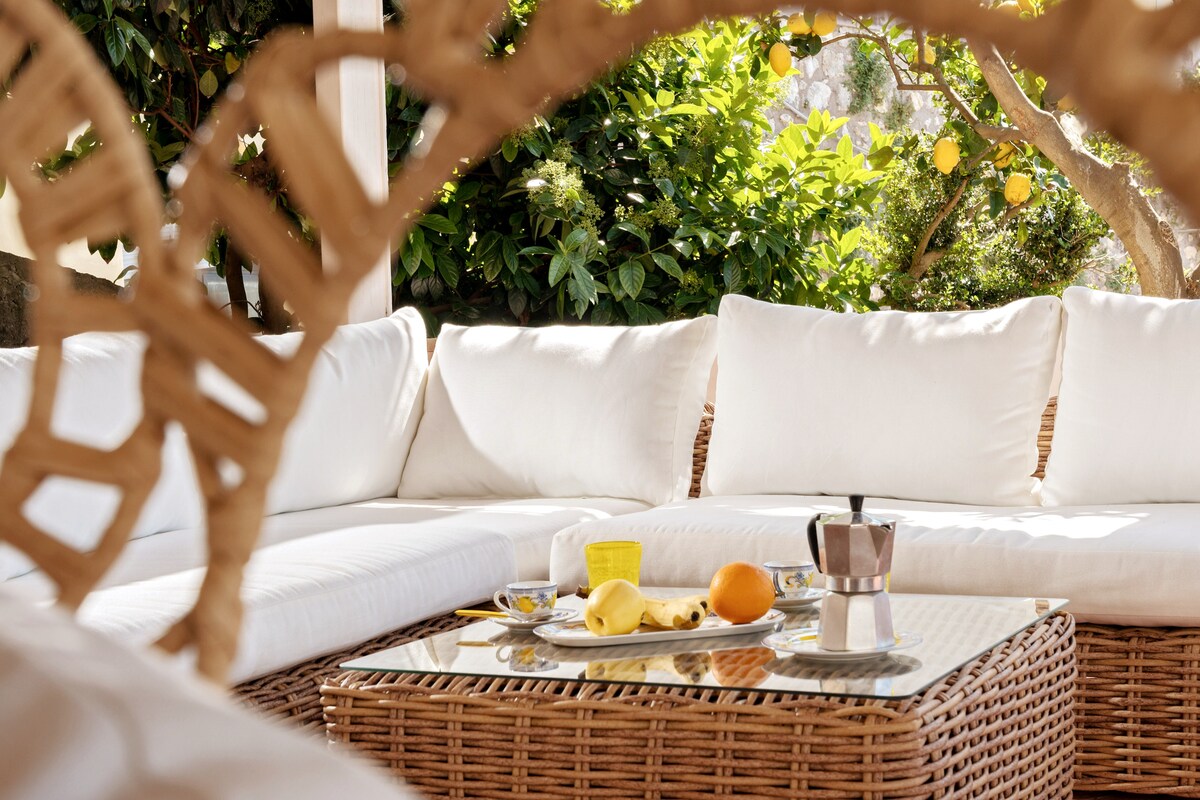 A lounge area features a spacious woven seating arrangement with white cushions, surrounded by greenery. A glass-topped table displays a variety of fruits and an Italian coffee maker. Natural light illuminates the area, creating a calm and inviting setting.