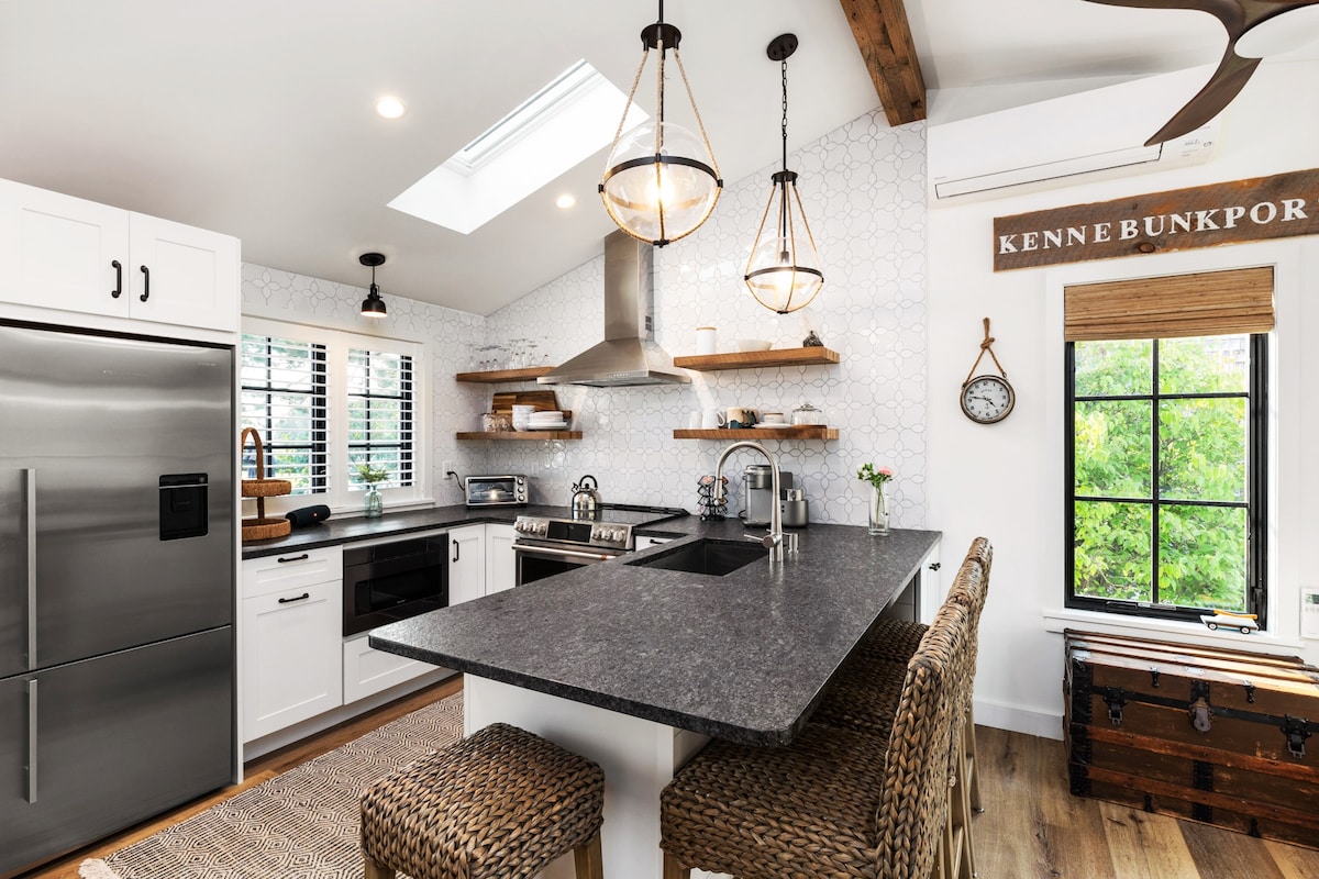 A modern kitchen features sleek appliances, including a stainless steel refrigerator and a gas range. Open shelving displays decorative items, while a spacious island with bar seating provides a gathering space. Natural light floods in through a skylight and window, accentuating the clean lines and neutral tones.