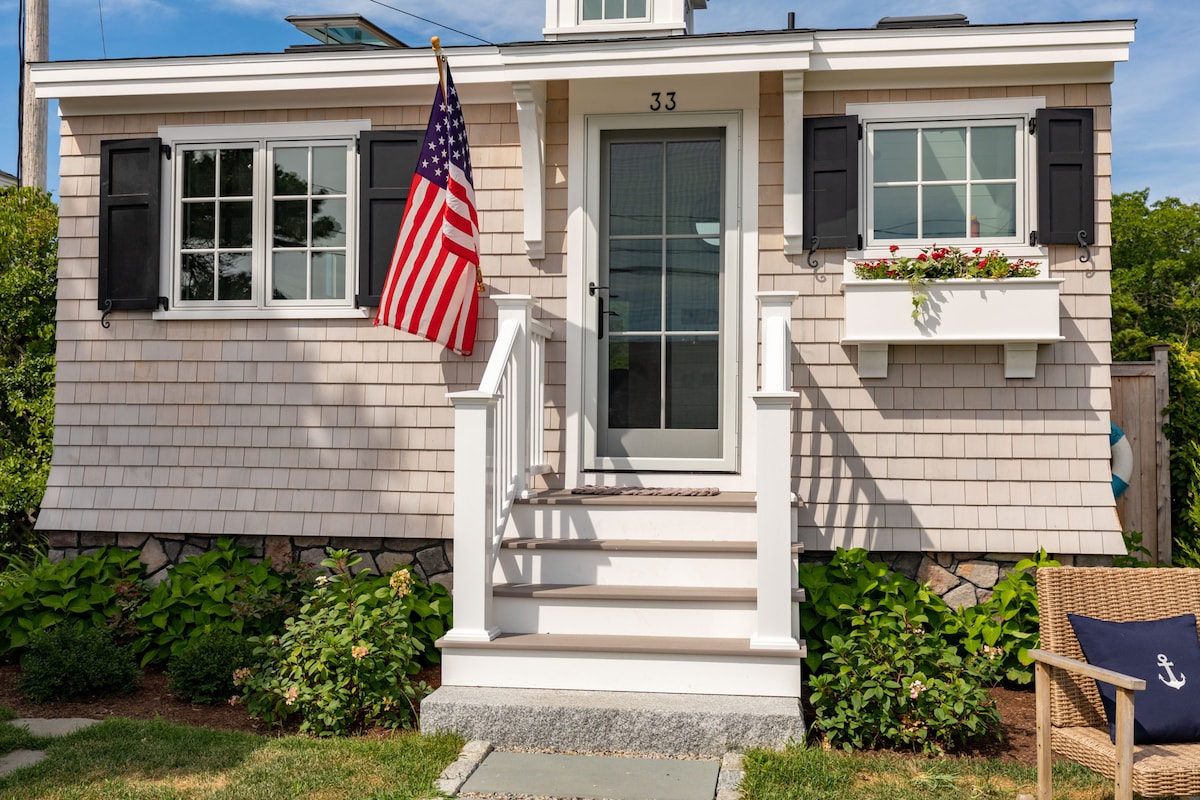 The exterior of a charming cottage is showcased with a light shingle facade and crisp white trim. A small staircase leads to a glass door framed by black shutters, while vibrant flowers bloom in the window box. An American flag hangs prominently at the entrance.