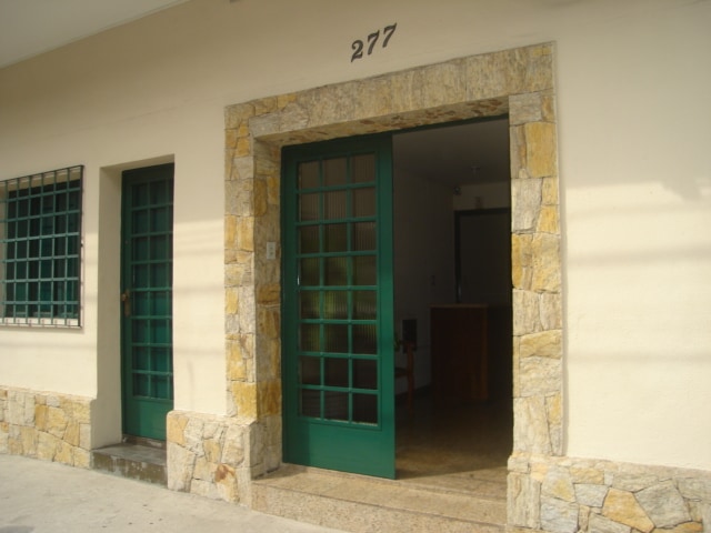 An exterior view of a building entrance features a stone facade with a vibrant green door and window frames. The door showcases paned glass, allowing natural light to filter in. Two matching doors are visible, framed by a neutral-colored wall.