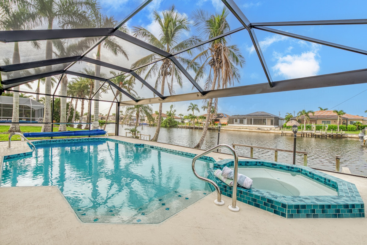 A newly resurfaced, tiled pool is featured within a screened enclosure, surrounded by palm trees. A jacuzzi is positioned adjacent to the pool, offering a calming water feature. The serene canal view is visible in the background, enhancing the outdoor space's inviting ambiance.