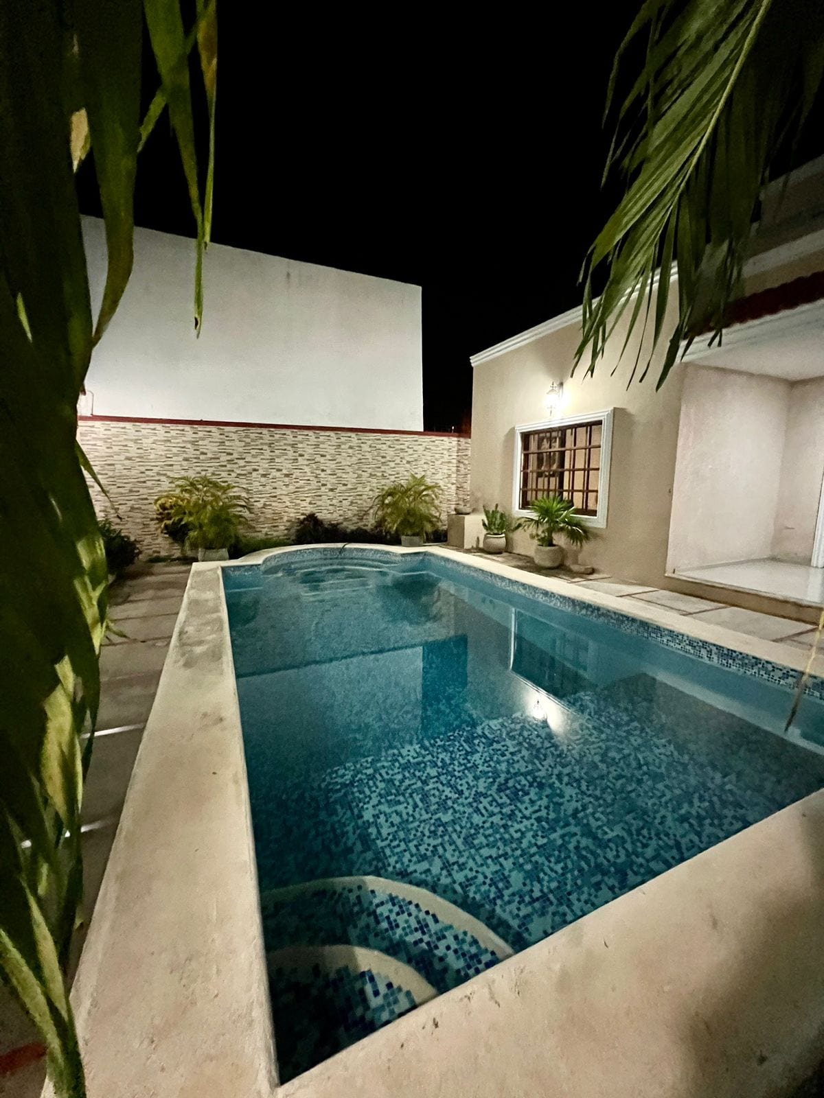 A swimming pool is surrounded by a tiled patio and bordered by lush greenery. The water reflects soft lighting from the nearby house, creating a serene evening atmosphere. Plants are placed along the edges, enhancing the private outdoor space.