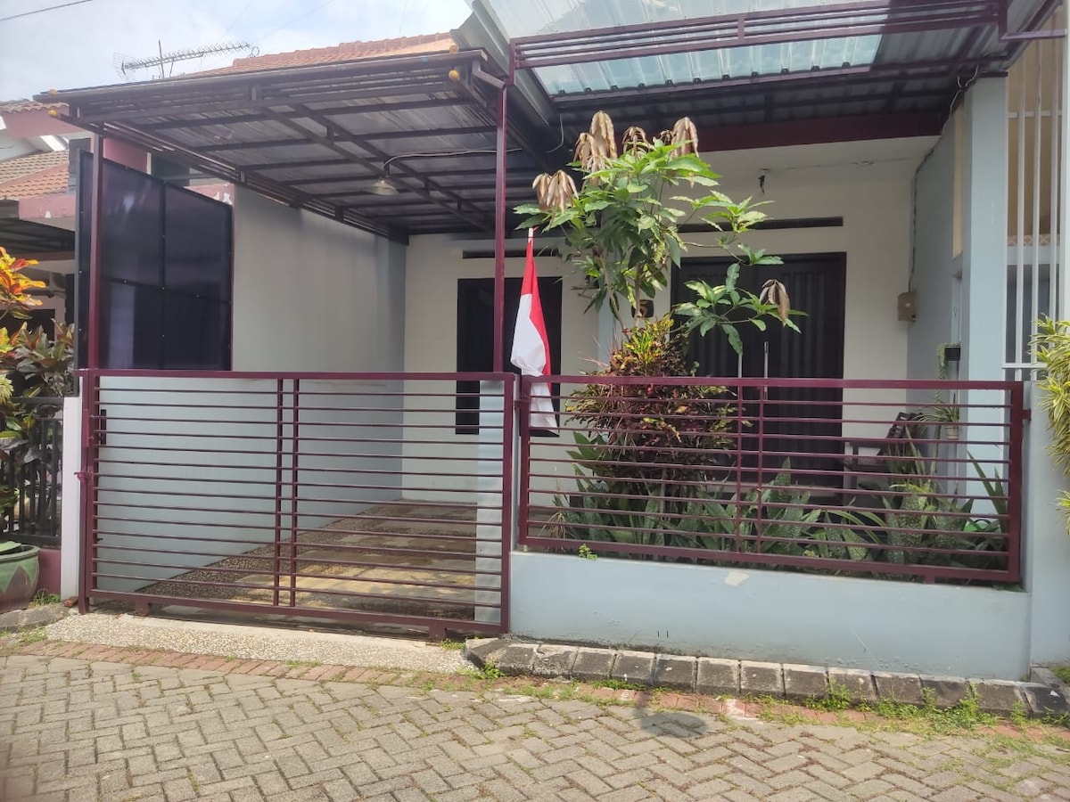 The entrance to the home is seen, featuring a covered porch with a few potted plants and a flag displayed prominently. The home is framed by a secure fence and a pathway leading up to the entryway, offering a welcoming approach.