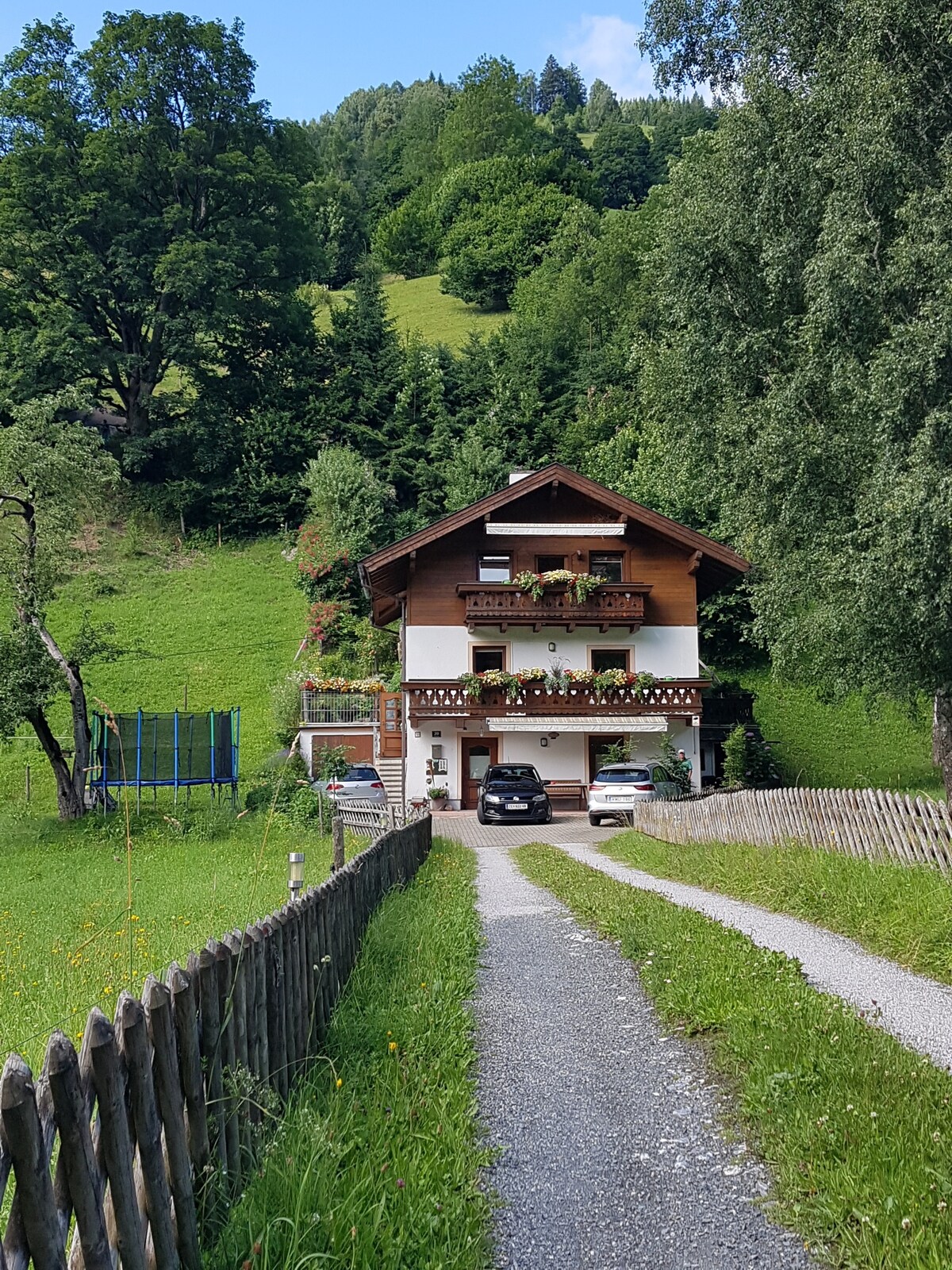 A charming two-story house is nestled in a green landscape, surrounded by trees and mountains. A gravel path leads up to the entrance, where flowers adorn the balconies. A trampoline is visible in the backyard, and two cars are parked in front of the house.