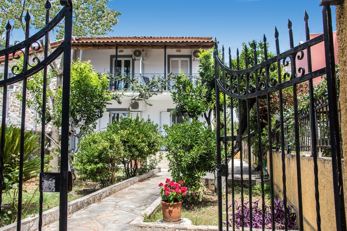 A wrought iron gate opens to a welcoming garden, framed by lush greenery and flowering plants. A stone pathway leads toward a two-story building, boasting a covered balcony adorned with blue shutters, hinting at comfortable accommodations indoors. The clear blue sky adds to the inviting atmosphere.