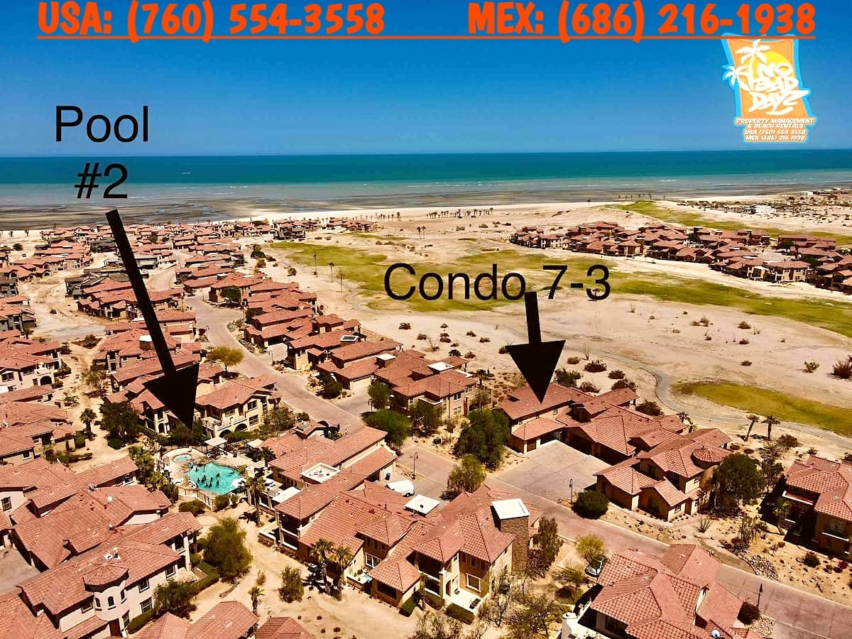 An aerial view showcases the layout of the community, highlighting the location of Condo 7-3 in proximity to communal Pool #2. The surrounding area features multiple residences with terracotta roofs and a vast expanse of sandy terrain leading to the shoreline.