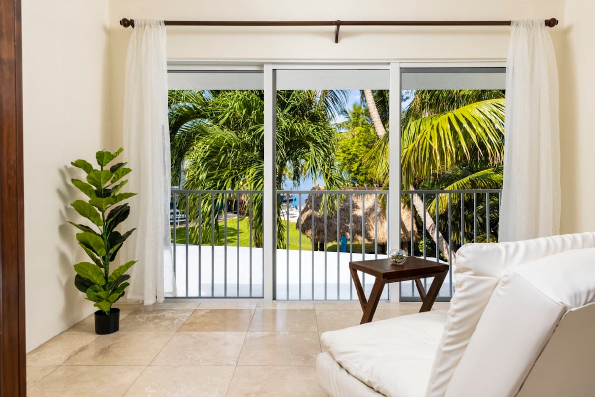 A serene lounge space features a white lounge chair and a small wooden table, with a potted green plant nearby. Floor-to-ceiling sliding glass doors offer a view of lush greenery and the blue waters of Florida Bay.