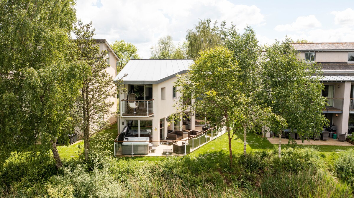 The exterior of Osprey Lodge is showcased, highlighting its modern design and landscaped surroundings. The large decking area is visible, furnished with outdoor seating, and framed by lush greenery. Glazed doors lead to the interior, blending indoor and outdoor living.