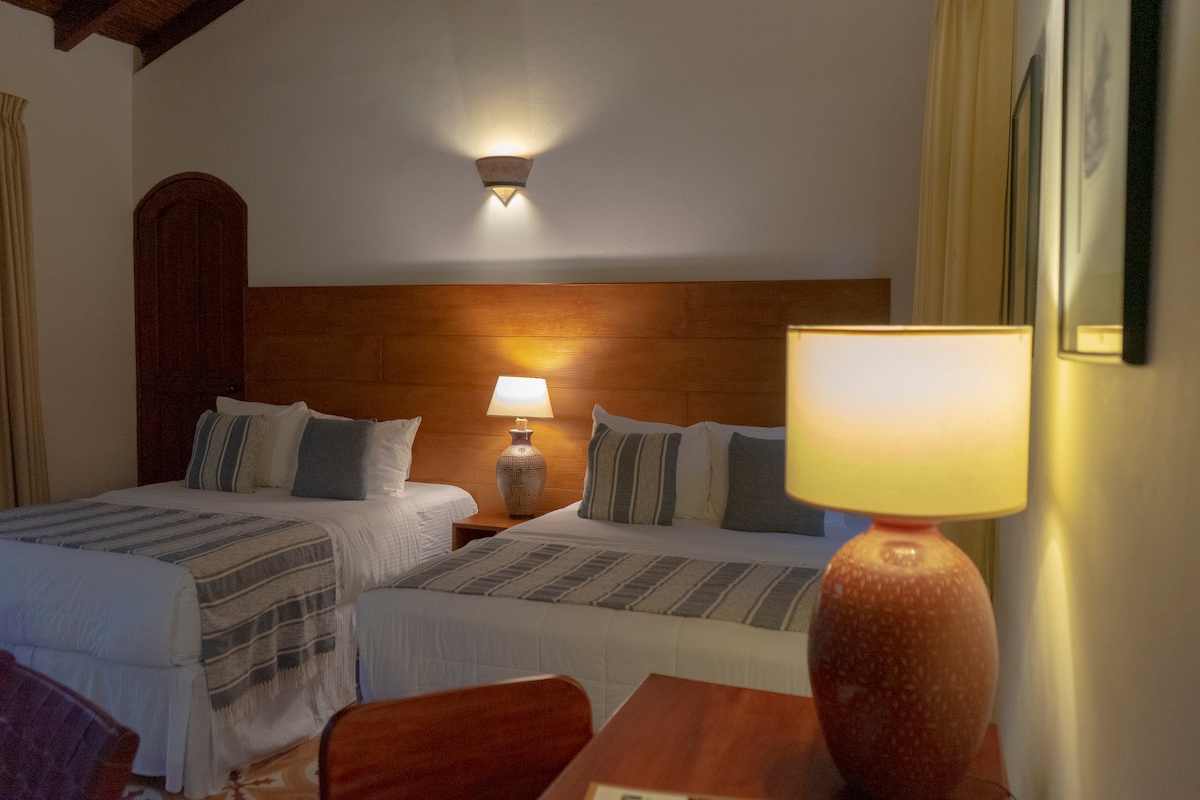 A serene bedroom features two spacious beds adorned with soft linens and decorative pillows. A bedside lamp sits on a wooden table, offering gentle illumination, while warm light reflects off the walls, enhancing the room's calming atmosphere. Natural light filters through the window, creating a cozy space.