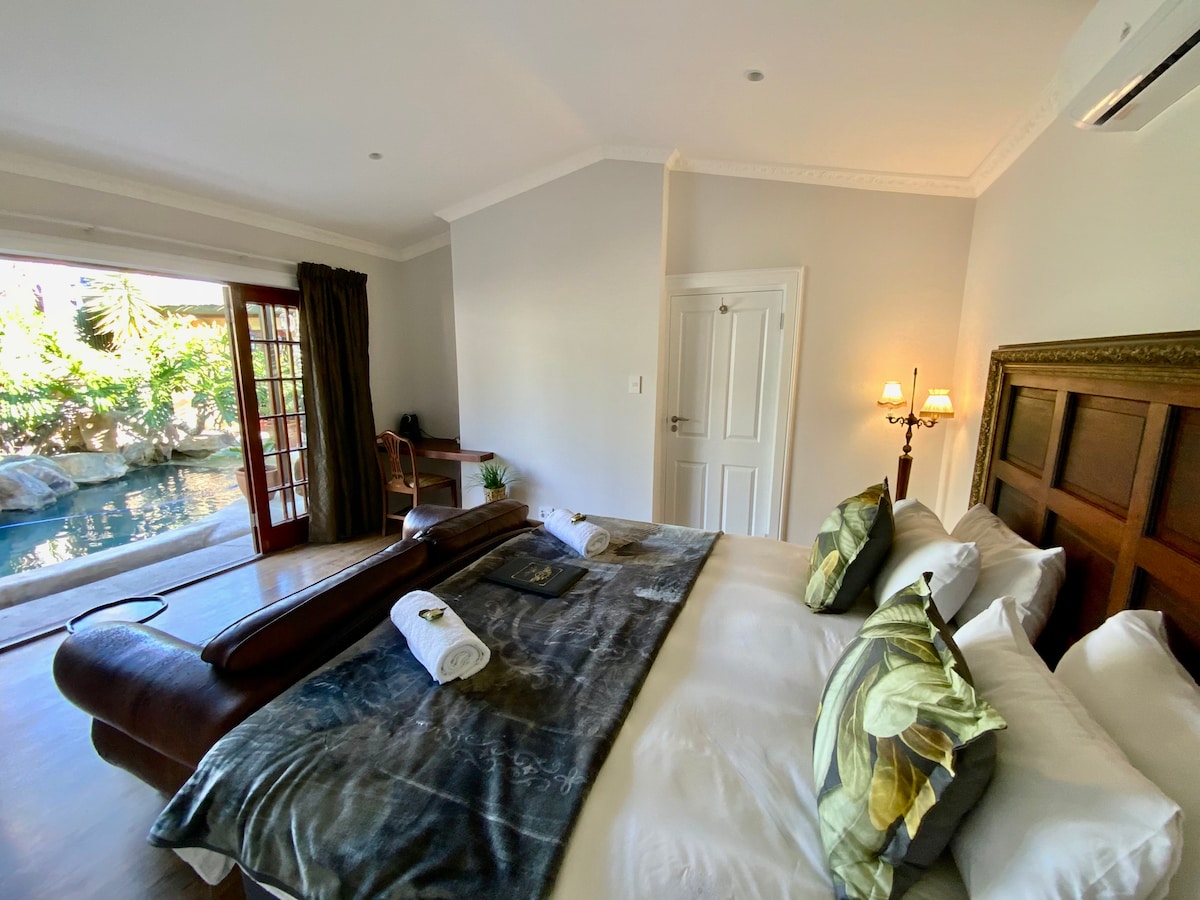 A spacious room is furnished with a large double bed, adorned with decorative pillows and a soft throw. French doors open to a tranquil pool area, complemented by natural light filtering in through the windows. A desk and elegant lamps enhance the serene atmosphere.