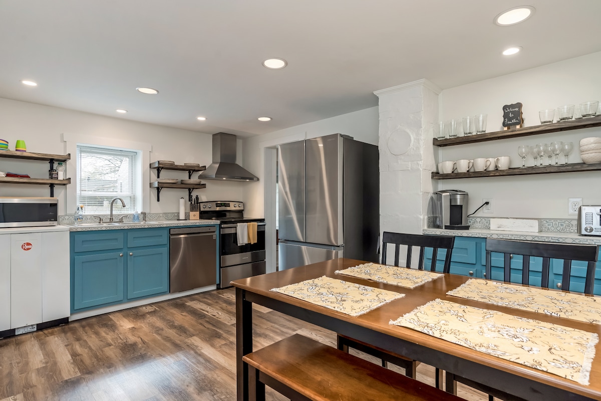 A modern kitchen features stainless steel appliances, including a refrigerator and oven. Light blue cabinetry creates a fresh atmosphere. A dining table is set for four, with decorative placemats. Open shelving displays glassware and kitchen essentials, while a window allows natural light to brighten the space.