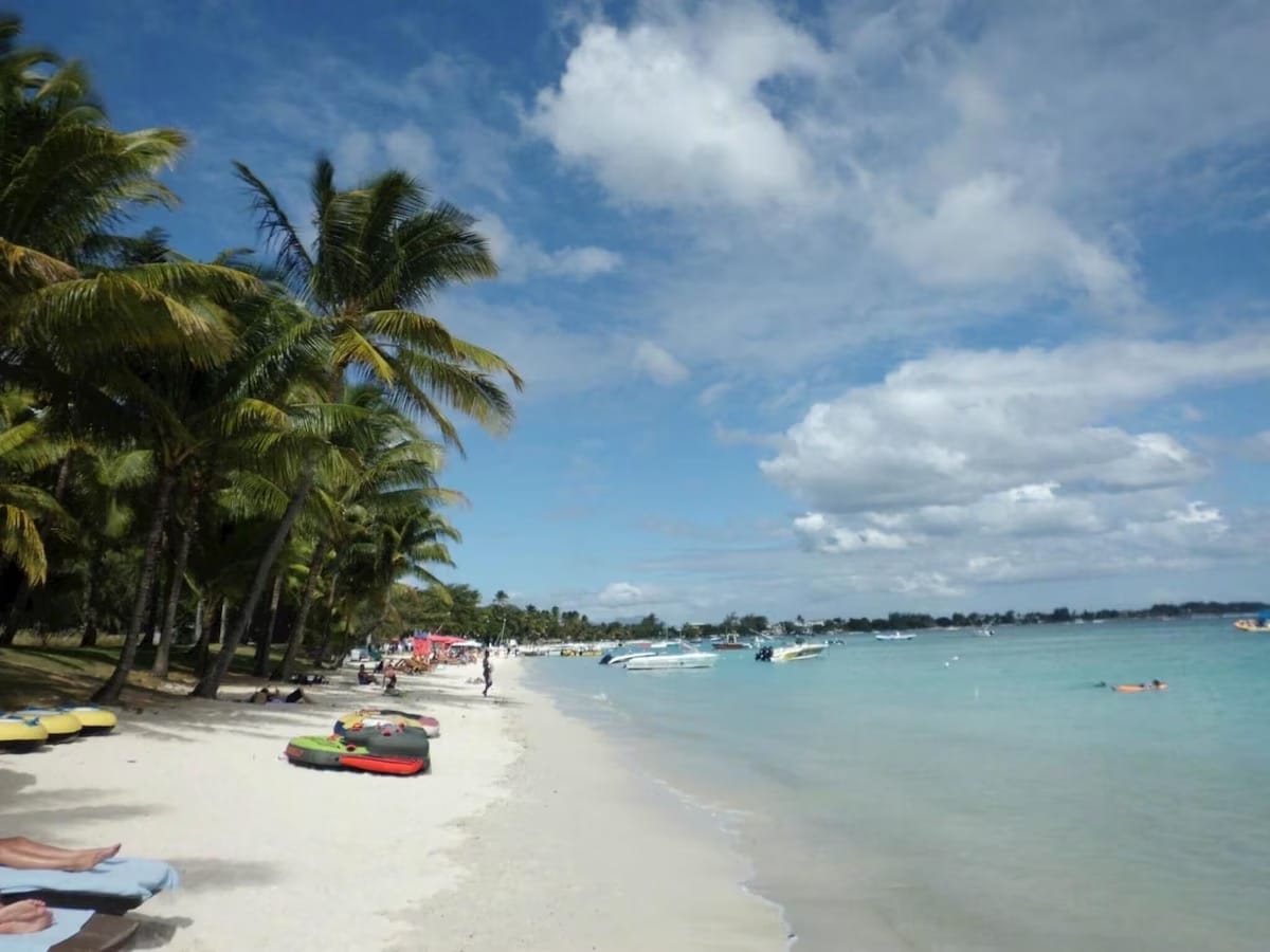 A sandy beach is lined with tall palm trees, creating a tropical atmosphere. Gentle waves lap at the shore, while several colorful beach mats are spread out on the sand. Small boats are seen in the distance, and people enjoy the serene beach environment.