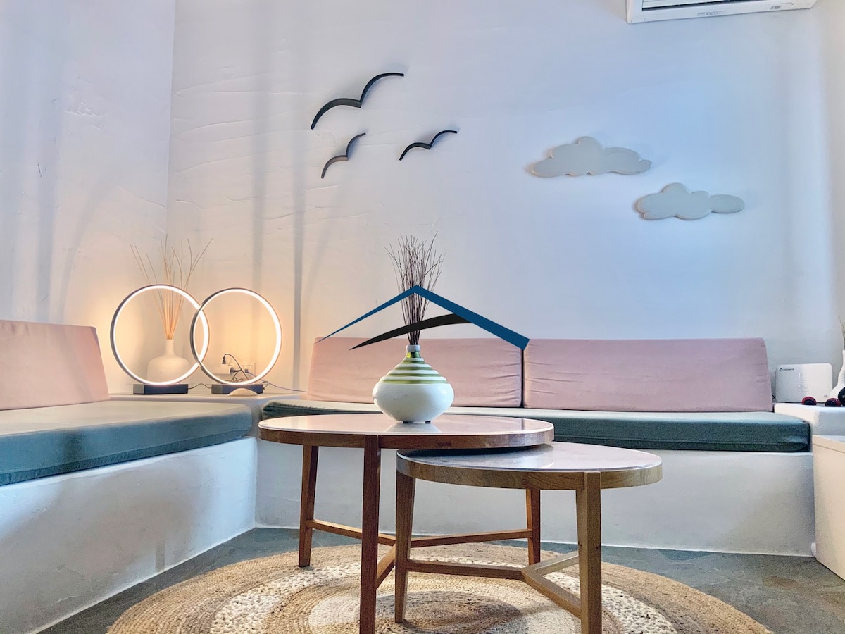 The living room area features built-in sofas with light pink cushions, accommodating a cozy seating arrangement. A circular decorative vase sits atop a wooden coffee table. Minimalist wall art in the shape of clouds and birds adds an airy feel to the space.