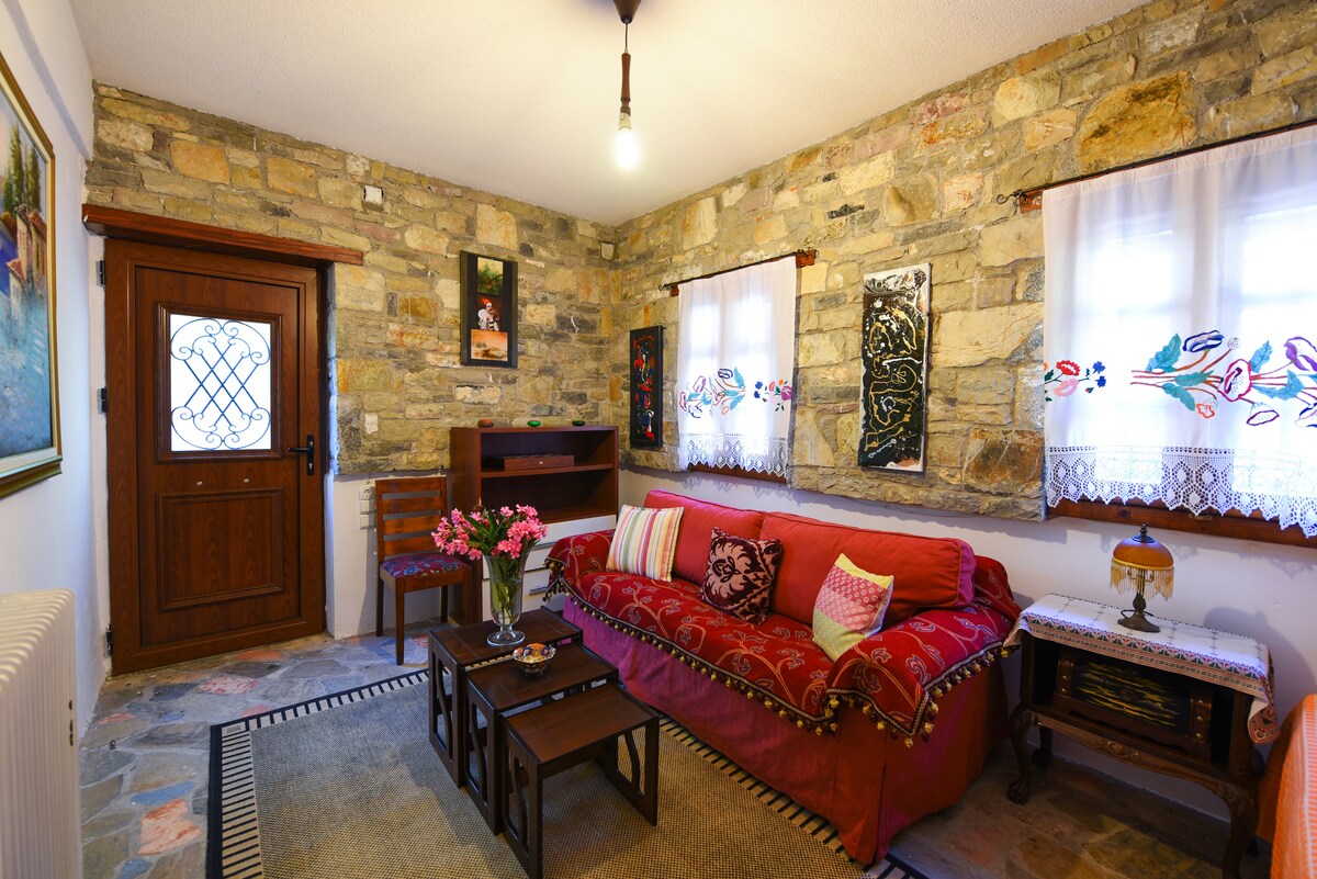 A cozy living area is presented with stone walls and wooden accents. A red sofa adorned with colorful cushions faces two dark wooden coffee tables. Natural light enters through a window with lace curtains, complementing the rustic decor and inviting atmosphere.