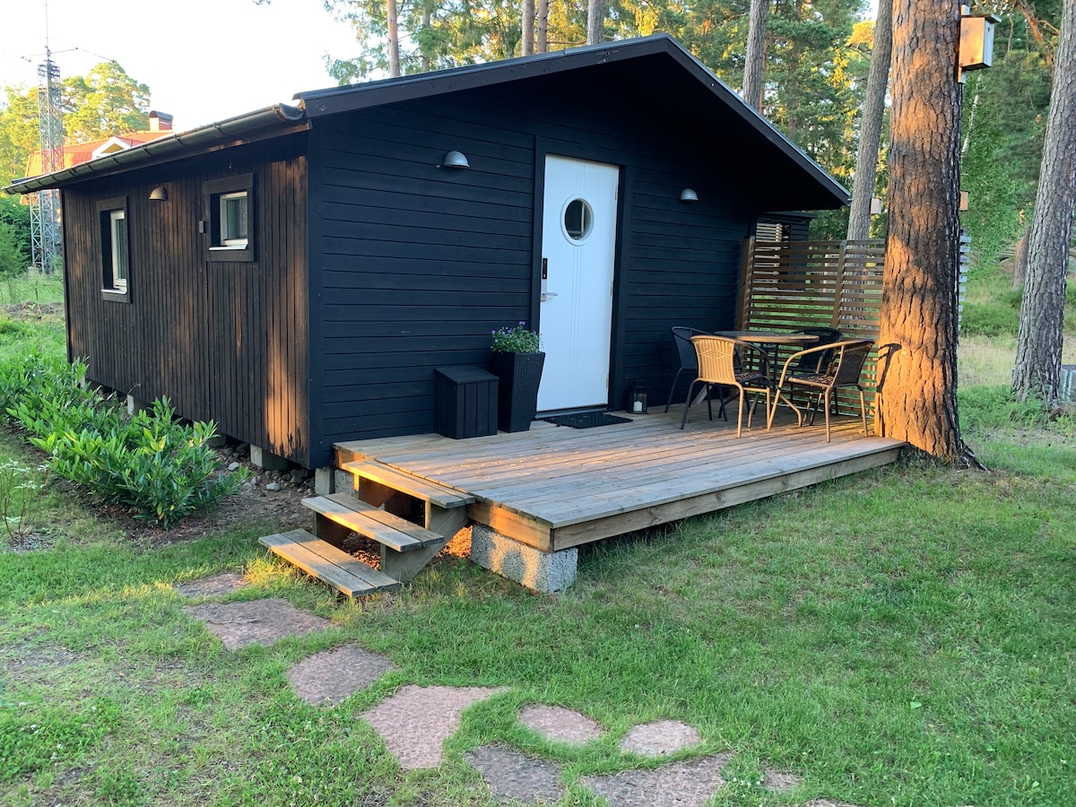 A small guesthouse is framed by lush greenery and trees, featuring a wood deck with steps leading up to the entrance. Outdoor seating arrangements are visible, accompanied by a potted plant near the door. The structure is painted in dark tones, creating a modern appearance.