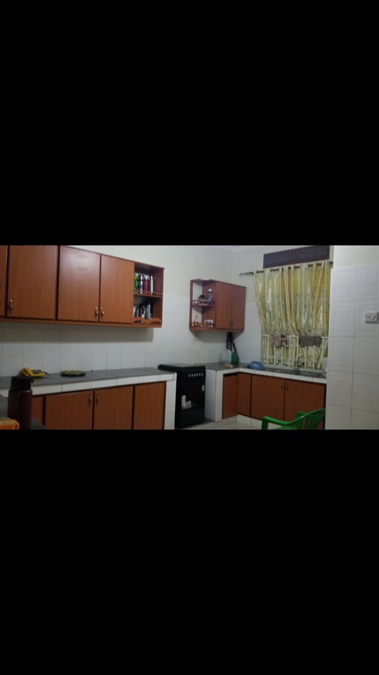 A simple kitchen space is presented with wooden cabinets and a small refrigerator. Countertops are clear, providing ample room for meal preparation. A window dressed with light curtains allows natural light to enter, enhancing the practicality of the space. A small table and green chair are visible.