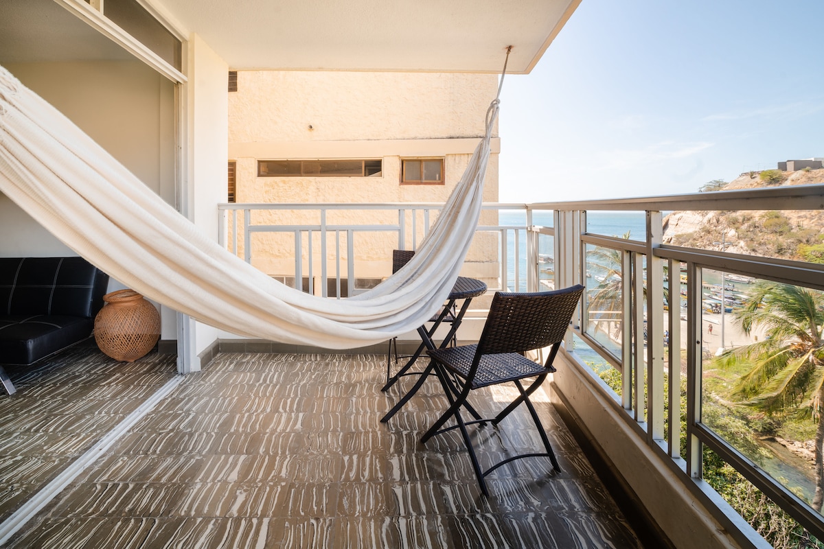 A spacious balcony features a hammock suspended for relaxation, complemented by two dark wicker chairs and a round decorative basket. The view includes the coastline and palm trees, creating a bright and airy atmosphere.