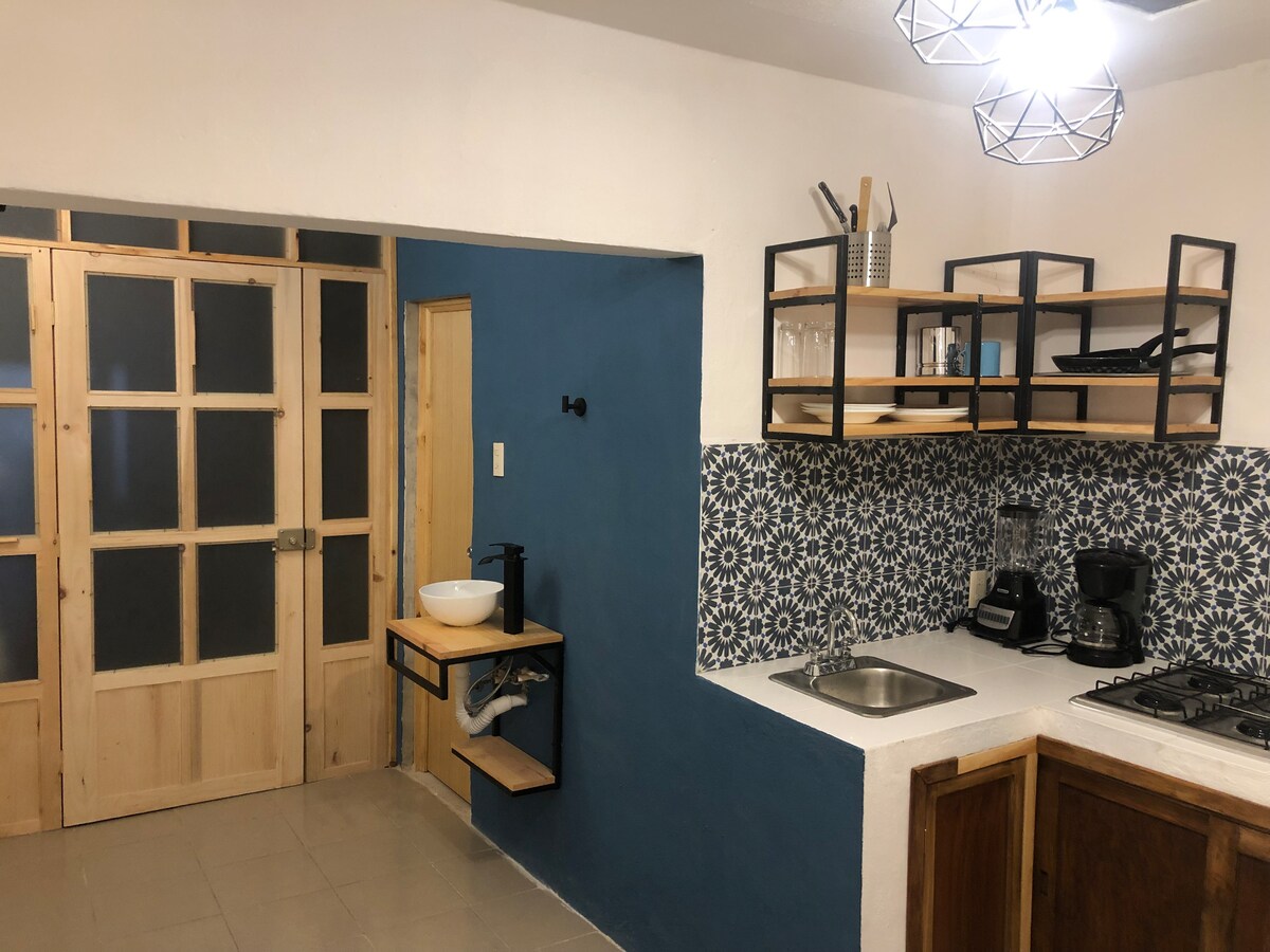 A modern kitchen area is featured, showcasing a blue accent wall and striking geometric shelving. Minimalist cabinetry complements the space, while a stainless steel sink and gas stove are prominently positioned. Built-in shelves display cooking essentials, enhancing the kitchen's functionality.