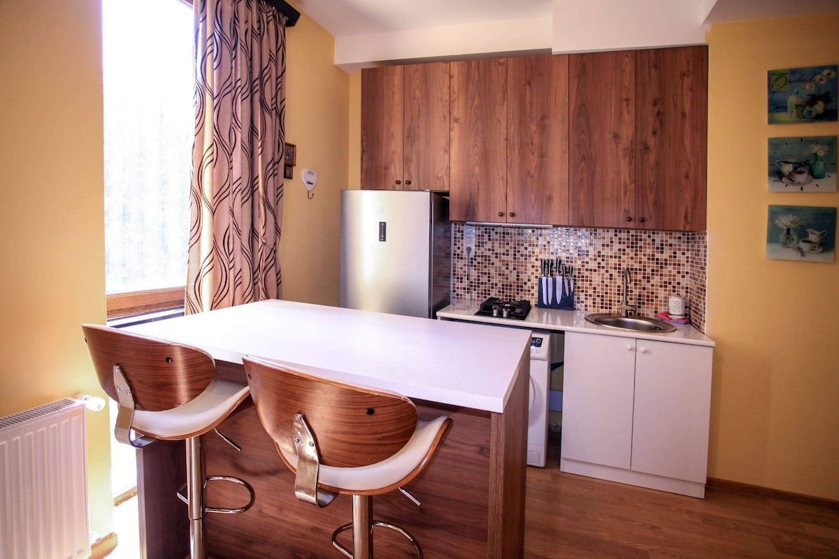 A modern kitchen area features warm wooden cabinets and a spacious island with two high stools. A refrigerator and a gas stove are visible, along with decorative tiles in the backsplash. Natural light enters through a large window, illuminating the space.