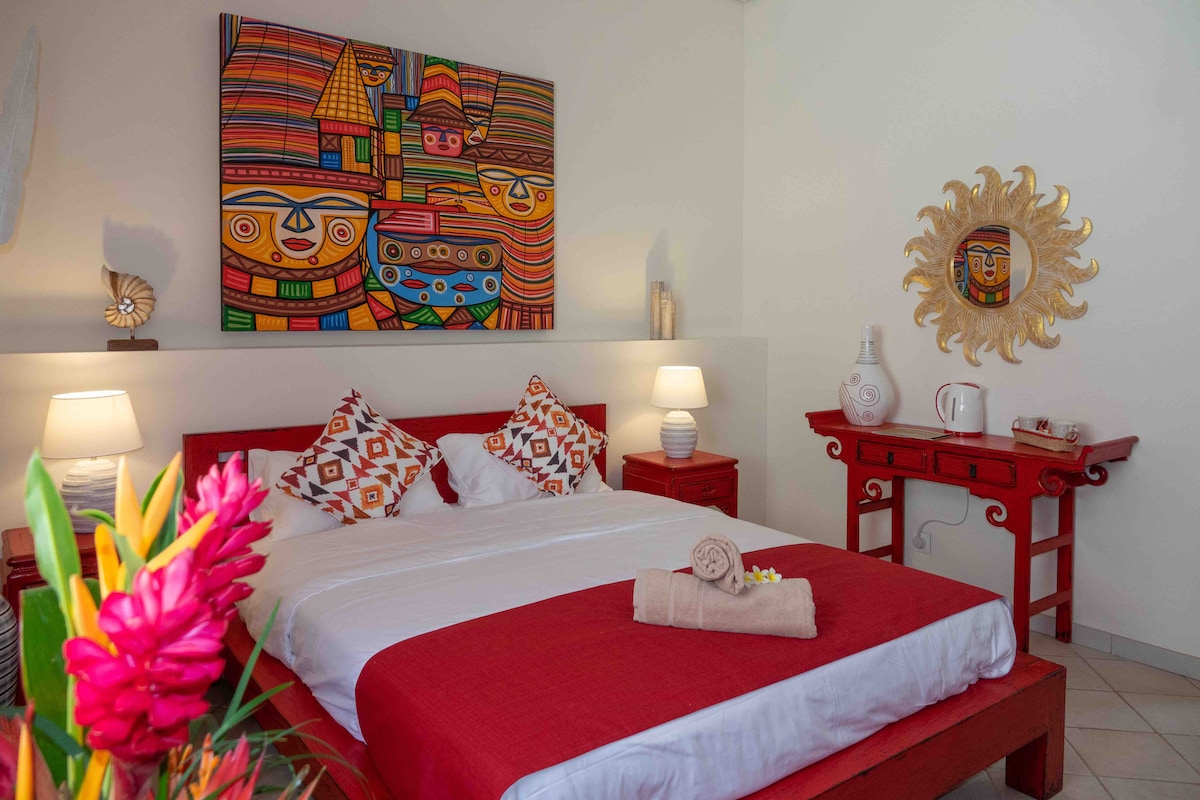 A well-appointed sleeping area features a spacious bed dressed in white linens and a vibrant red bedspread. Decorative pillows add a splash of color. Artistic wall decor and a sun-shaped sculpture enhance the ambiance, with soft lighting provided by bedside lamps.