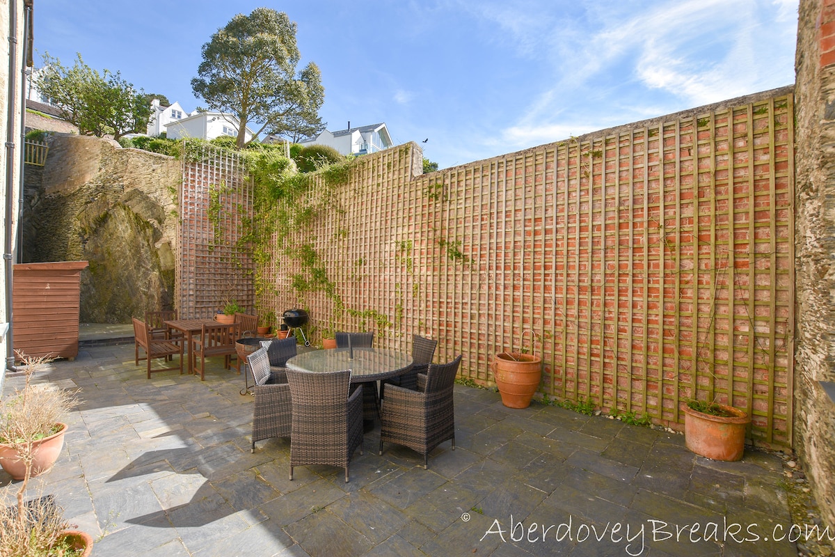 A private courtyard featuring outdoor furniture, including a round table and several chairs. Potted plants line the perimeter, and natural stone flooring extends throughout the area. The surrounding brick walls are adorned with greenery, creating a secluded outdoor retreat.