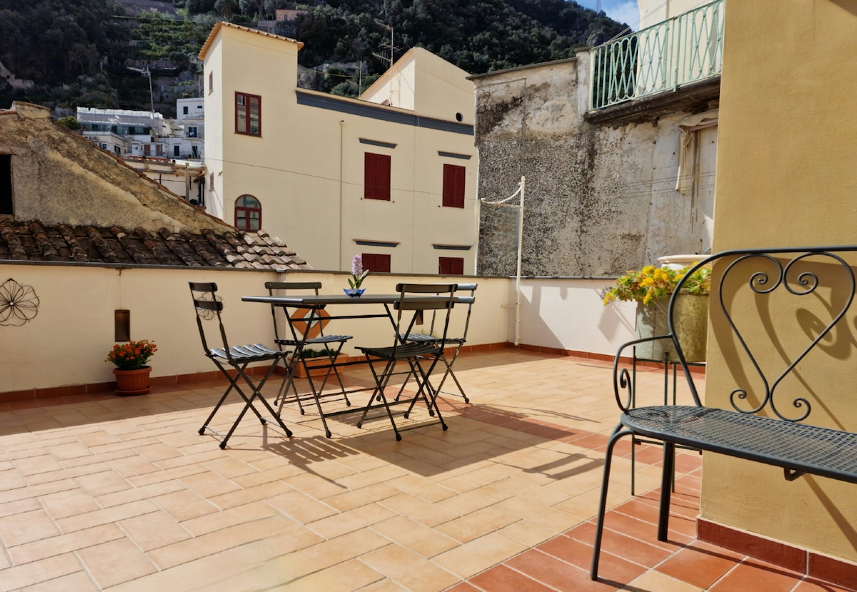 A communal terrace features a table and four folding chairs on terracotta tiles, offering a space for relaxation. Potted flowers add a touch of color, while surrounding buildings with red window shutters create a charming urban backdrop. Sunlight illuminates the area.
